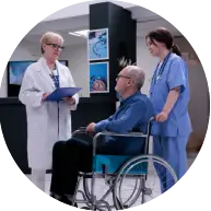 A doctor in a lab coat discusses with a patient in a wheelchair, accompanied by a nurse in scrubs, in a hospital setting.